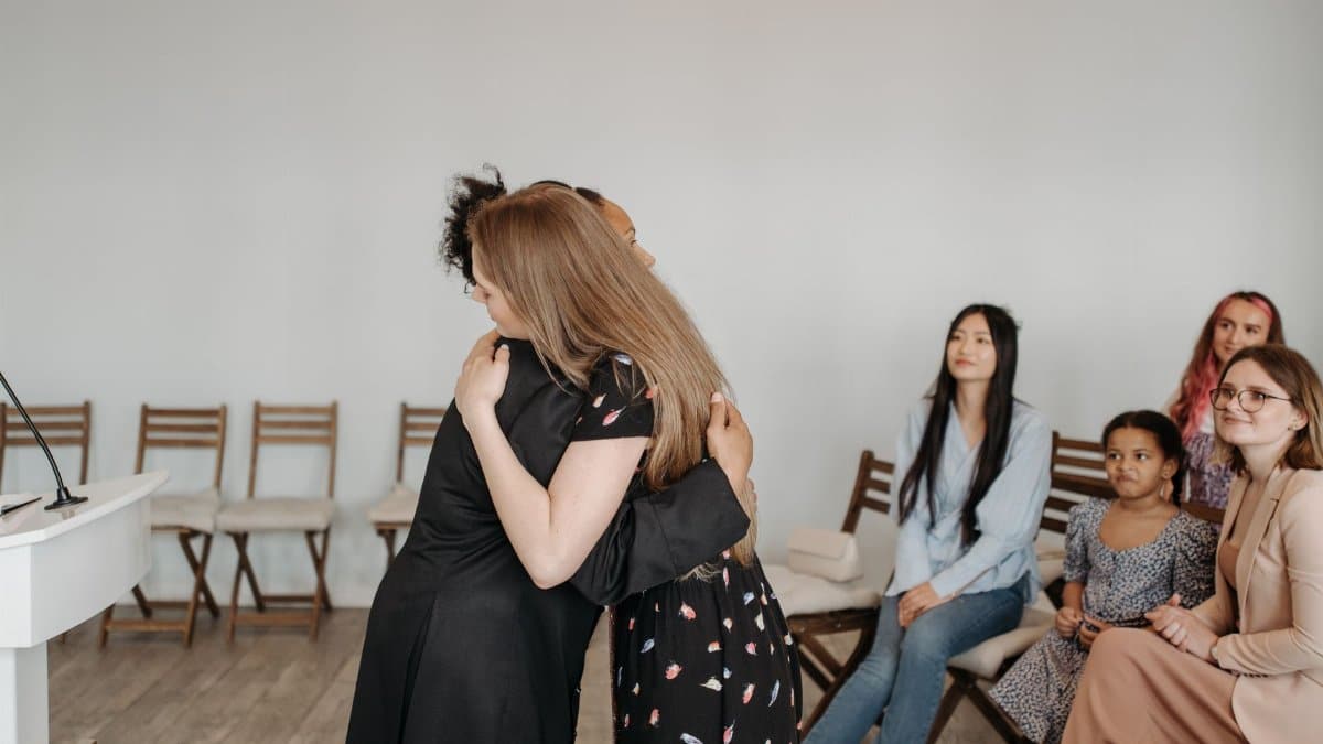 Diverse group of women sharing an embrace indoors while others observe. A sense of community and support is depicted.