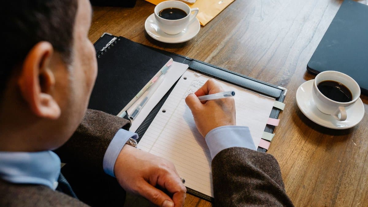 Crop unrecognizable man in formal clothes sitting at table with cup of coffee and writing notes in notepad during business meeting