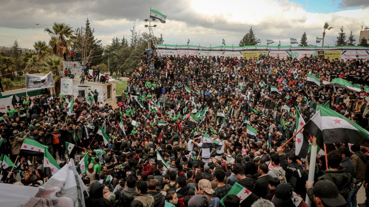 Large crowd gathers in Idlib for a protest, waving Syrian flags and expressing activism.