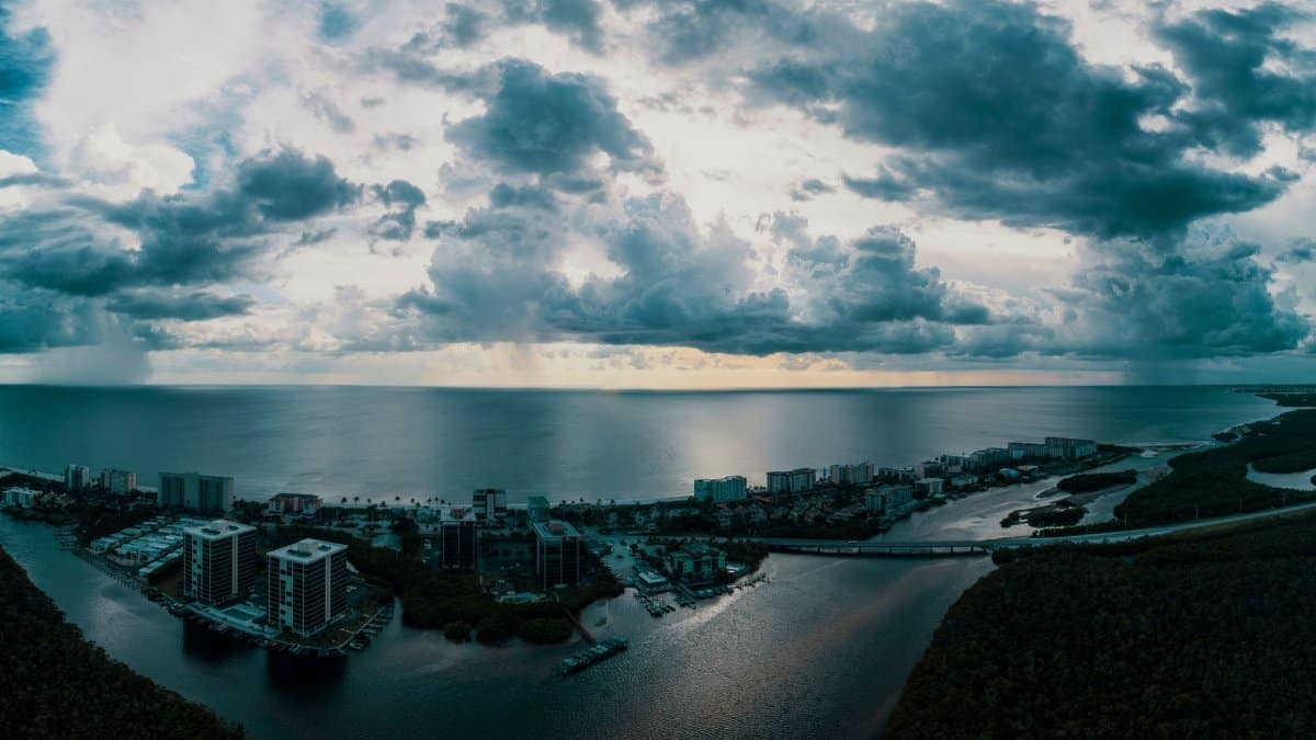A breathtaking aerial view of a coastal city under dramatic cloudscape, showcasing buildings, ocean, and horizon.