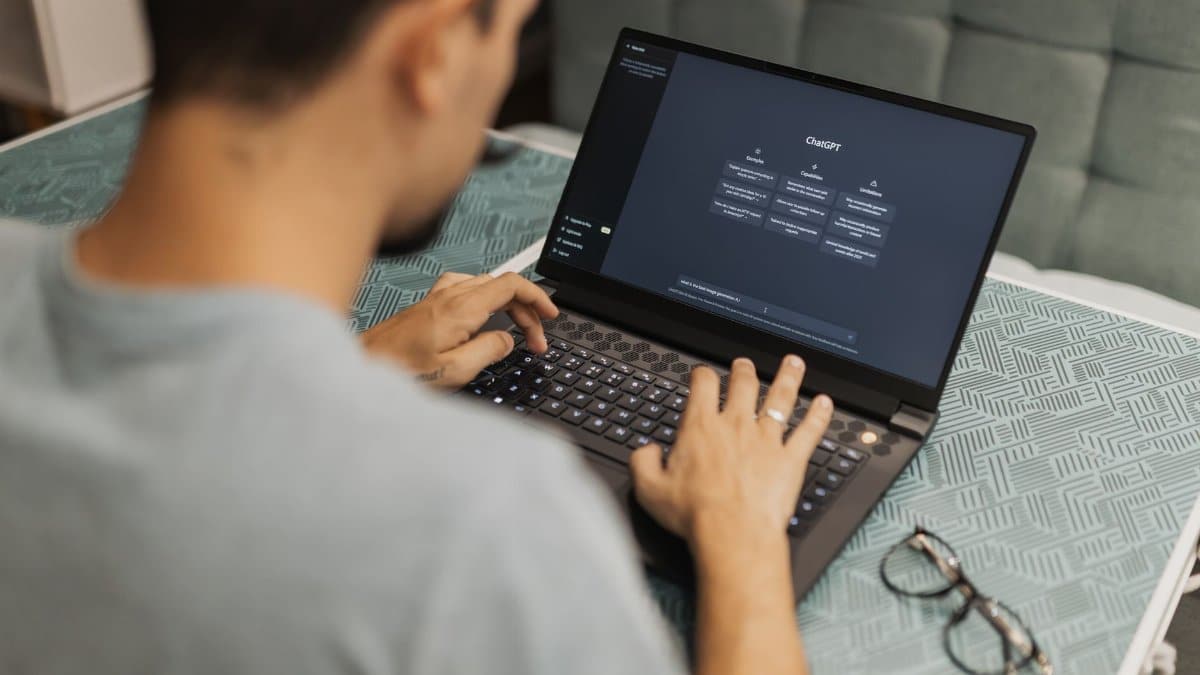 Man using laptop with AI interface, typing with focused attention. Indoors with eyewear beside.