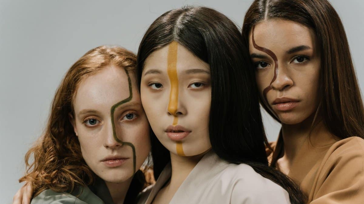Artistic portrait of three diverse women with unique face paint, expressing creativity and diversity in a studio setting.