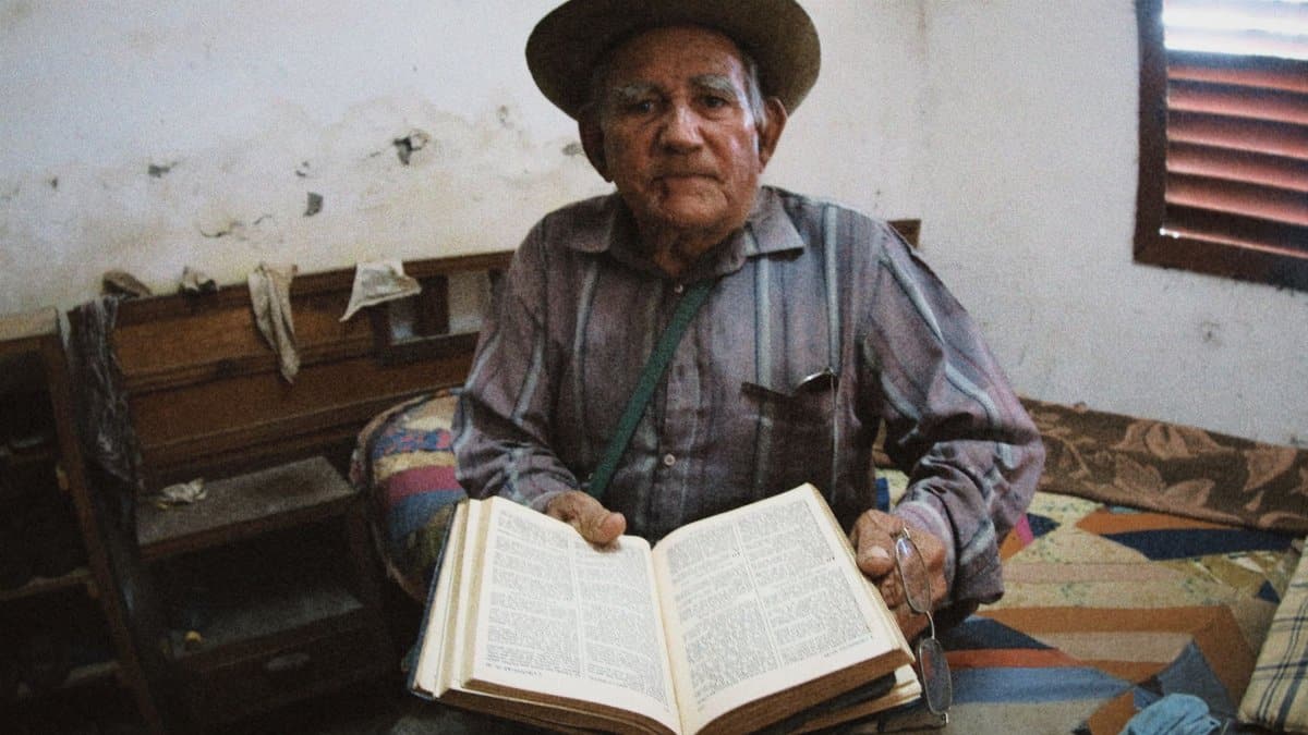 Elderly man in a rustic room reading a large book, depicting wisdom and solitude.