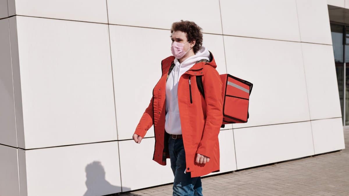 Delivery worker in red jacket and mask walking outdoors with thermal bag, practicing safety measures.