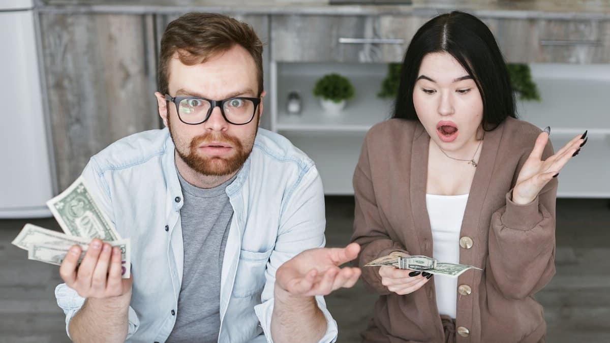 Interracial couple shocked while handling cash in a modern kitchen. Captivating expressions indoors.