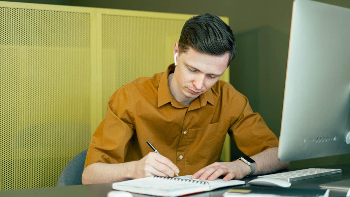 A man concentrating on his studies, taking notes at a desk with a computer.