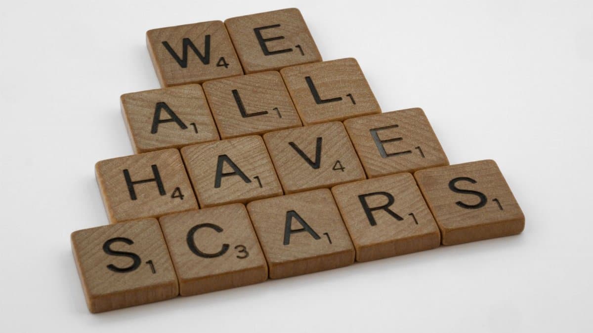 Wooden tiles spell 'We All Have Scars' on a white background in pyramid form.