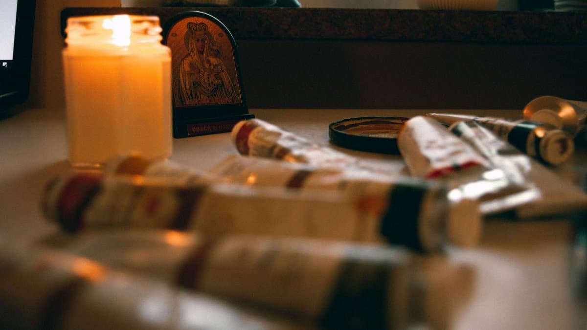 A warm, cozy workspace featuring a candlelit table with art supplies and a religious icon.