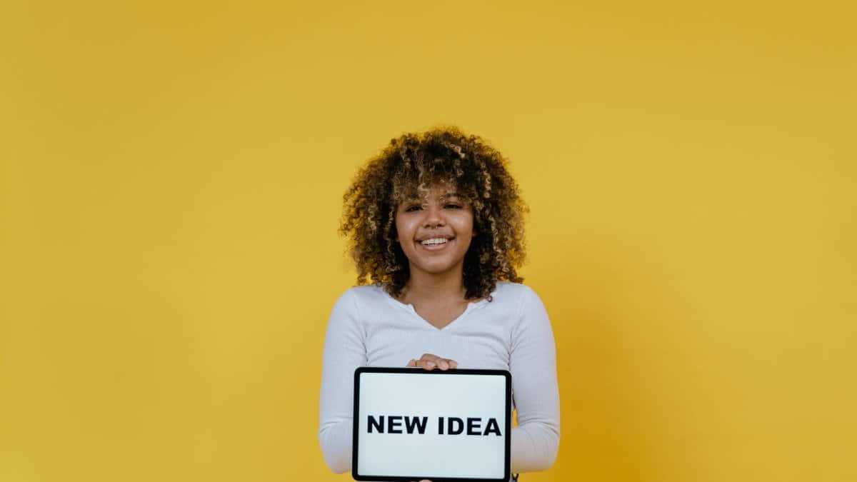 Young woman in white holding a tablet displaying 'New Idea' against a yellow background.