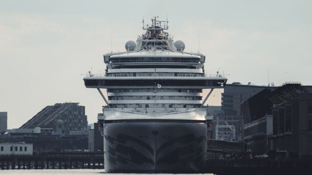 A massive cruise ship docked at an urban harbor, amidst tall buildings and clear skies.