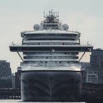 A massive cruise ship docked at an urban harbor, amidst tall buildings and clear skies.
