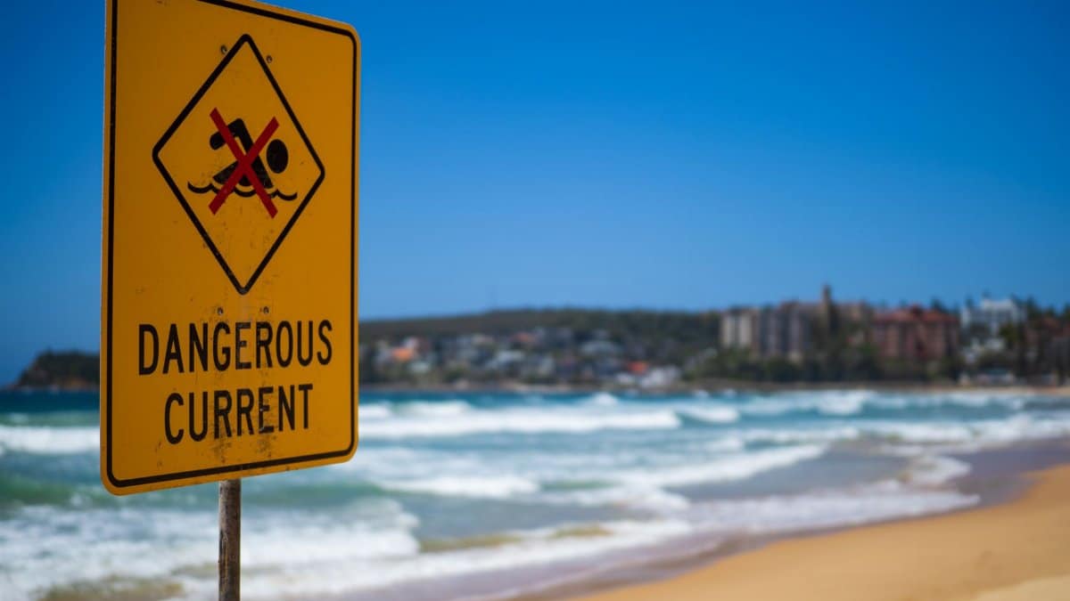 Dangerous current warning sign on Manly Beach in Australia, with blue sky and ocean waves.