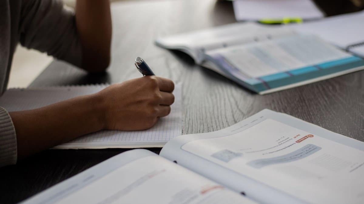 Side view of unrecognizable African American student sitting at table with textbooks while taking note in copybook in classroom with anonymous classmates on blurred background