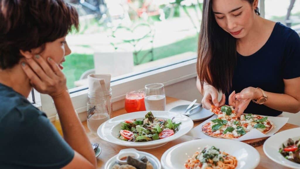 Content Asian woman taking slice of pizza while sitting at table near friend with delicious salad and pasta in restaurant