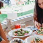 Content Asian woman taking slice of pizza while sitting at table near friend with delicious salad and pasta in restaurant