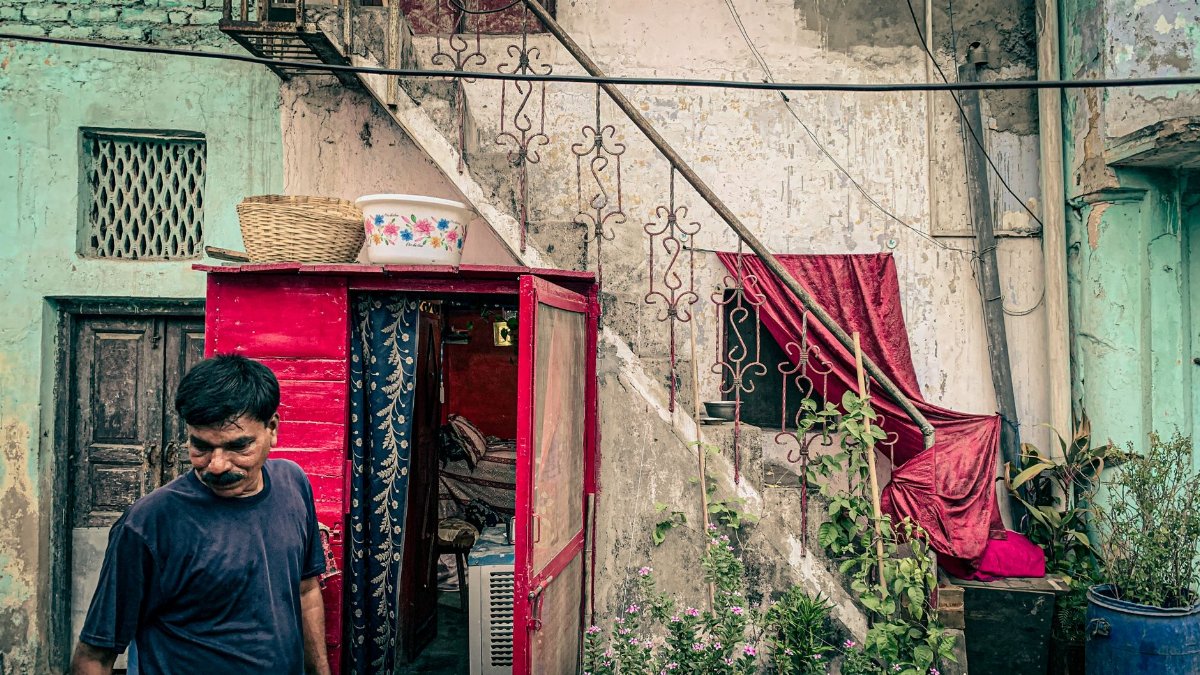 A man stands in front of a rustic building with vibrant red elements, offering a glimpse into urban decay and lifestyle.