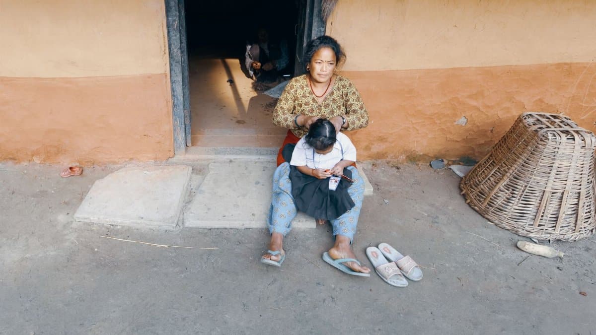A mother braids her daughter's hair outside their rural home, capturing candid family life.