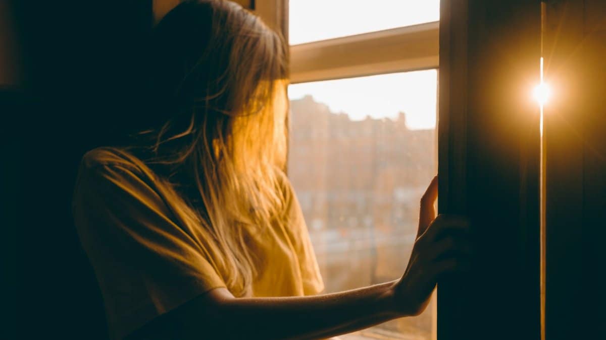 A woman stands at a window in Dublin during sunset, basking in warm sunlight.