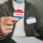 A person in a mask holding an 'I Voted' sticker, emphasizing civic engagement.