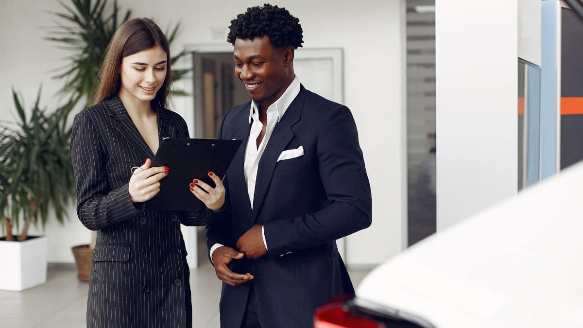 Cheerful multiethnic stylish female agent and smiling customer in formal suit discussing contract details while standing in modern car showroom in daylight