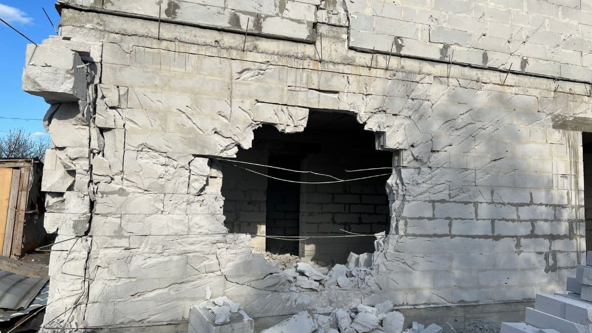 Concrete building wall with a large hole, showing damage and rubble, under a clear blue sky.