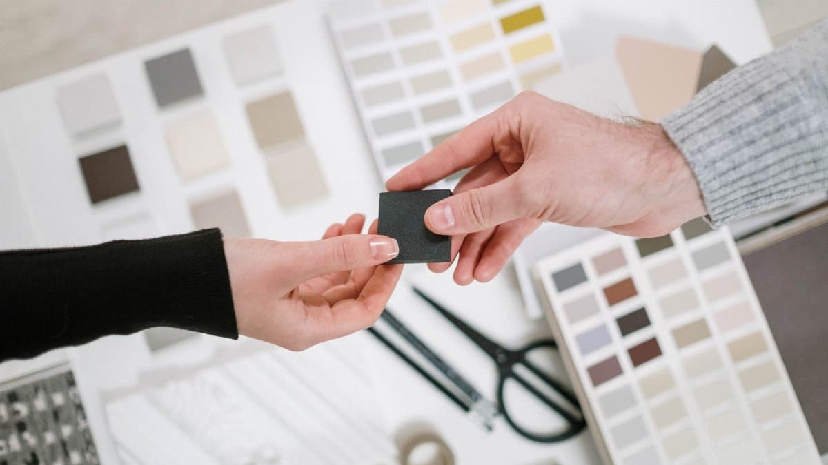 Two hands exchange a color sample during a collaborative design meeting with various palettes.