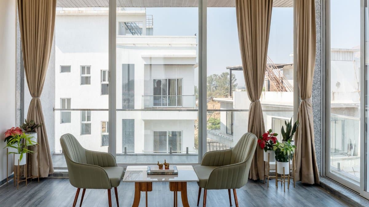 A spacious and modern living room with large windows, elegant chairs, and lush greenery.