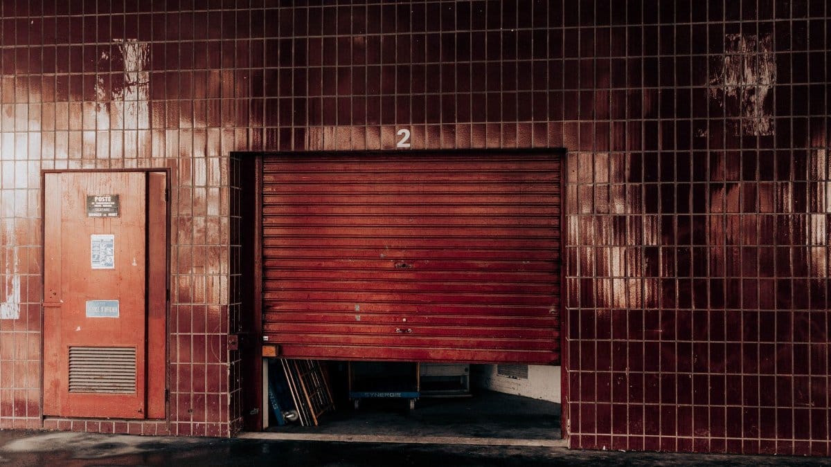 Aged garage exterior with security shutter and door between tiled wall with number on sunny day