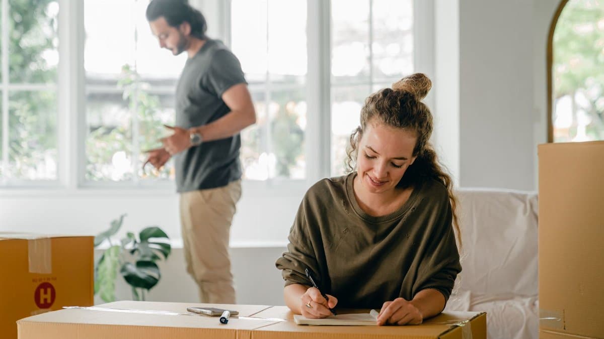 Positive young woman writing in notepad while sitting against cardboard boxes during relocation in new house with husband