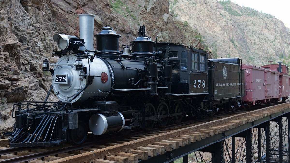 A historic steam locomotive crossing a railway bridge in a rocky canyon setting.