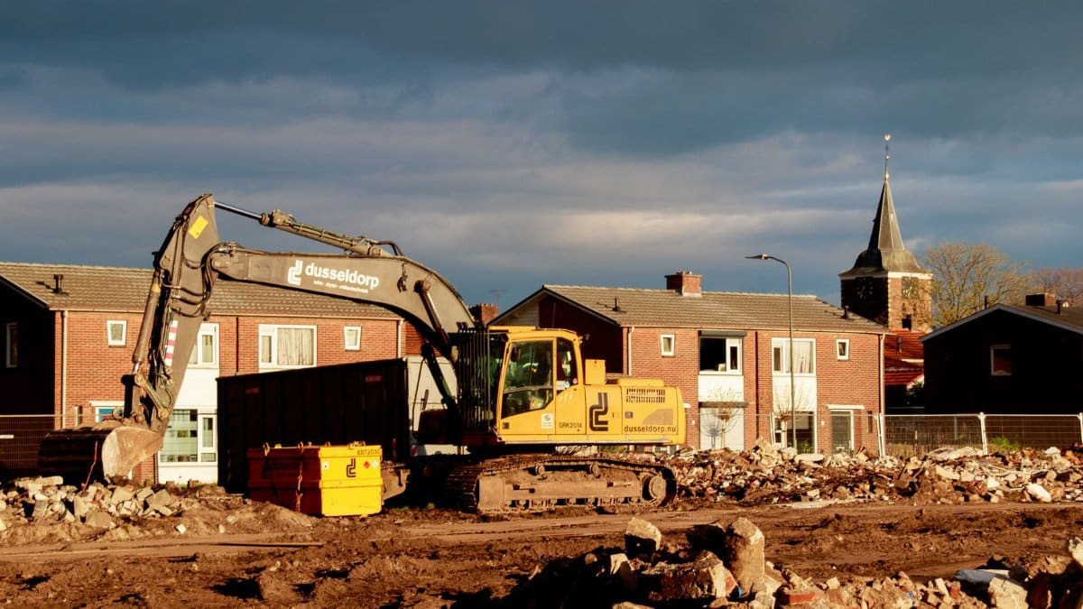 Excavator at a construction site near residential houses under a dramatic sky.
