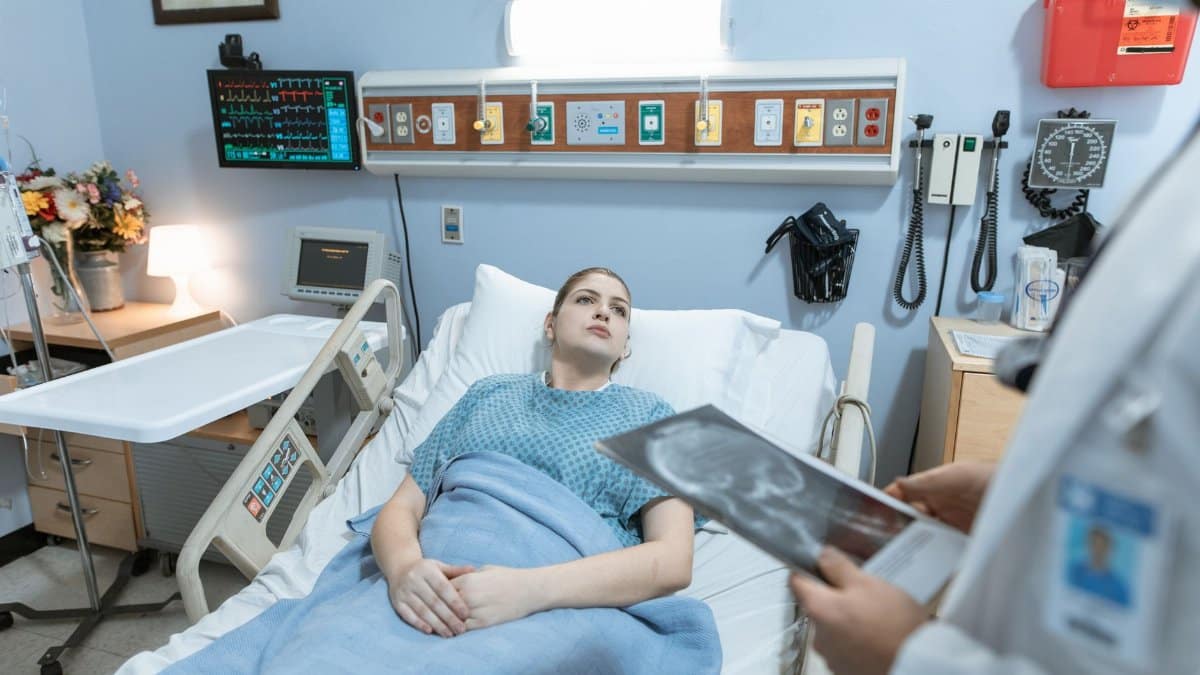 A female patient in a hospital gown receives medical attention from a healthcare professional holding an x-ray in a hospital ward.