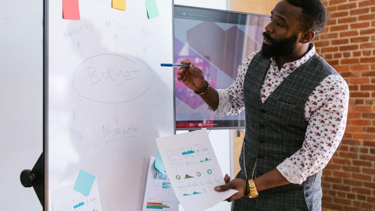 A businessman explains budget strategies using a whiteboard in an office setting.