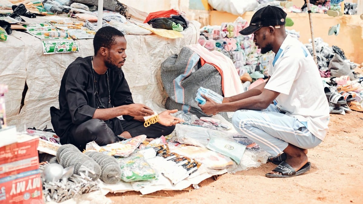 Two men engaging in a marketplace deal outdoors with various goods on display.