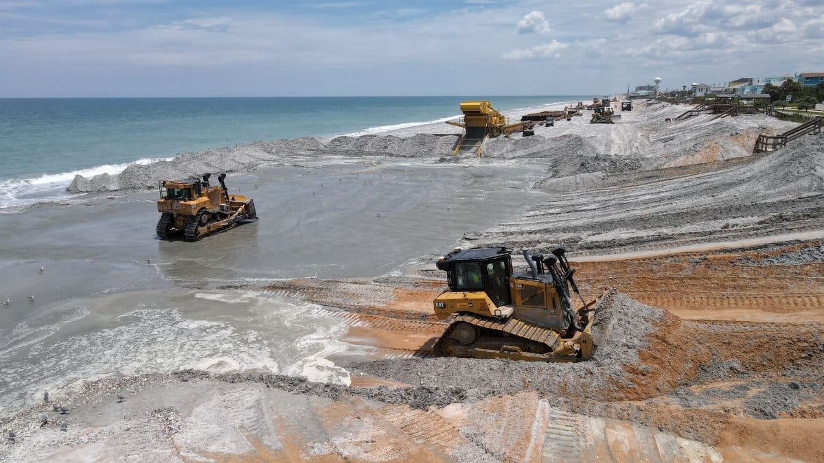 Heavy bulldozers reshape a coastline for beach restoration work under clear skies.