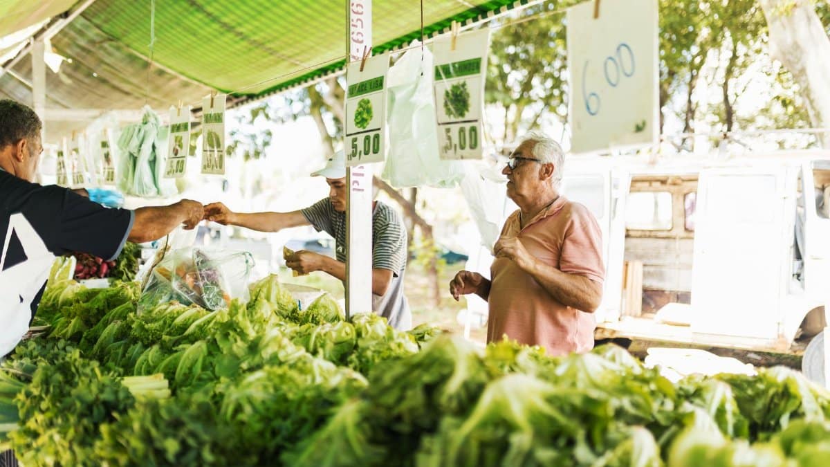 Customers purchasing fresh vegetables at an outdoor farmers market under green canopy.
