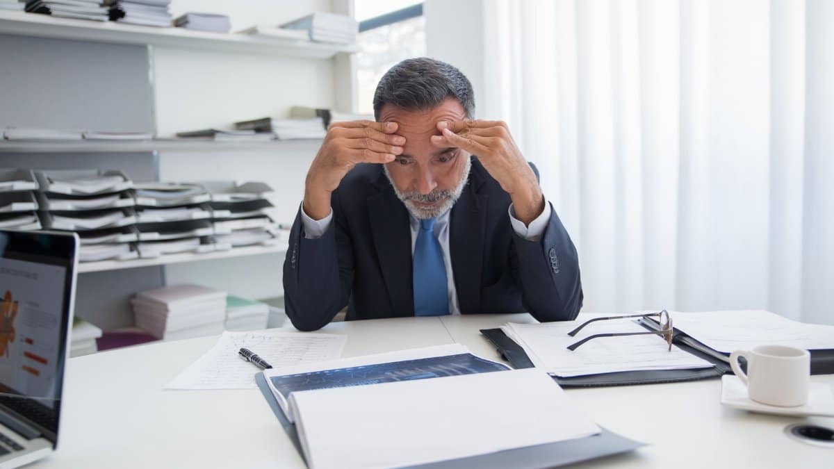 An elderly businessman in a suit appears stressed while sitting at a desk with documents in an office.