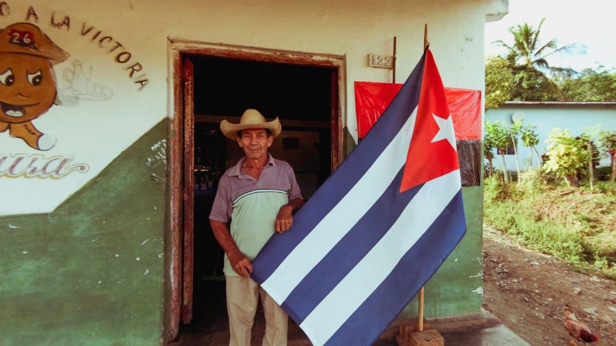 Elderly man in a straw hat proudly holds a Cuban flag outside a rustic house in a rural setting.