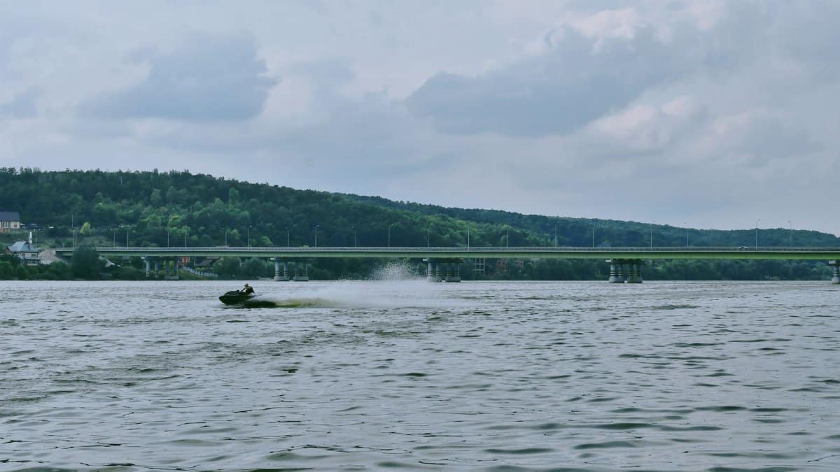 A person enjoying a jet ski ride on a wide calm river beneath a long bridge, at daytime.