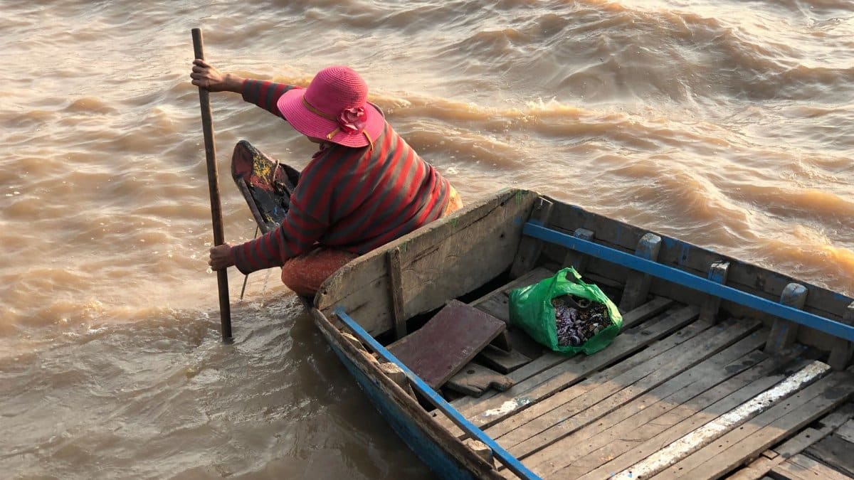 A woman in a striped shirt and pink hat rows a wooden boat on muddy water.