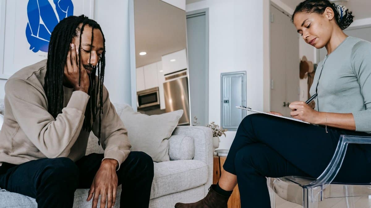 Attentive African American female with clipboard taking notes near sad male partner touching face on couch at home