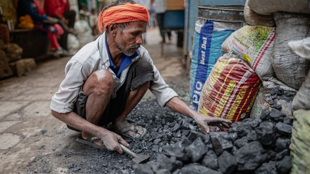 An Indian street worker wearing an orange turban sorts coal in Varanasi, highlighting daily life in a bustling market.