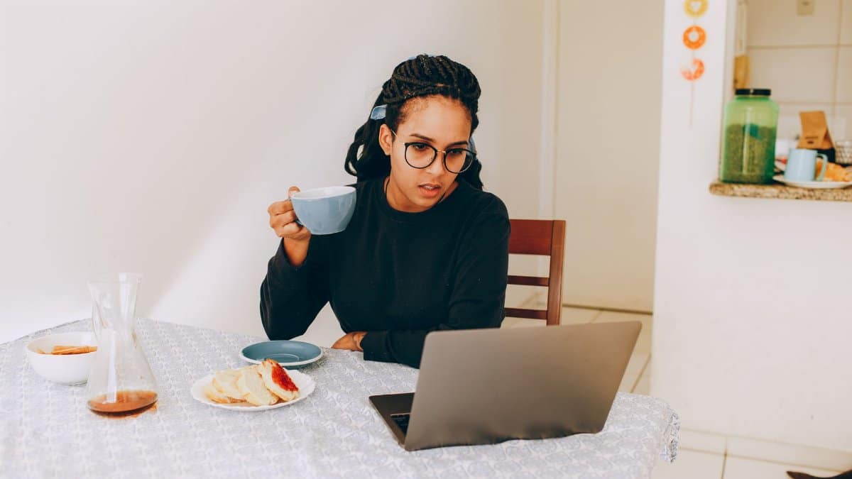 Young woman enjoying coffee while working remotely on her laptop at home kitchen table.