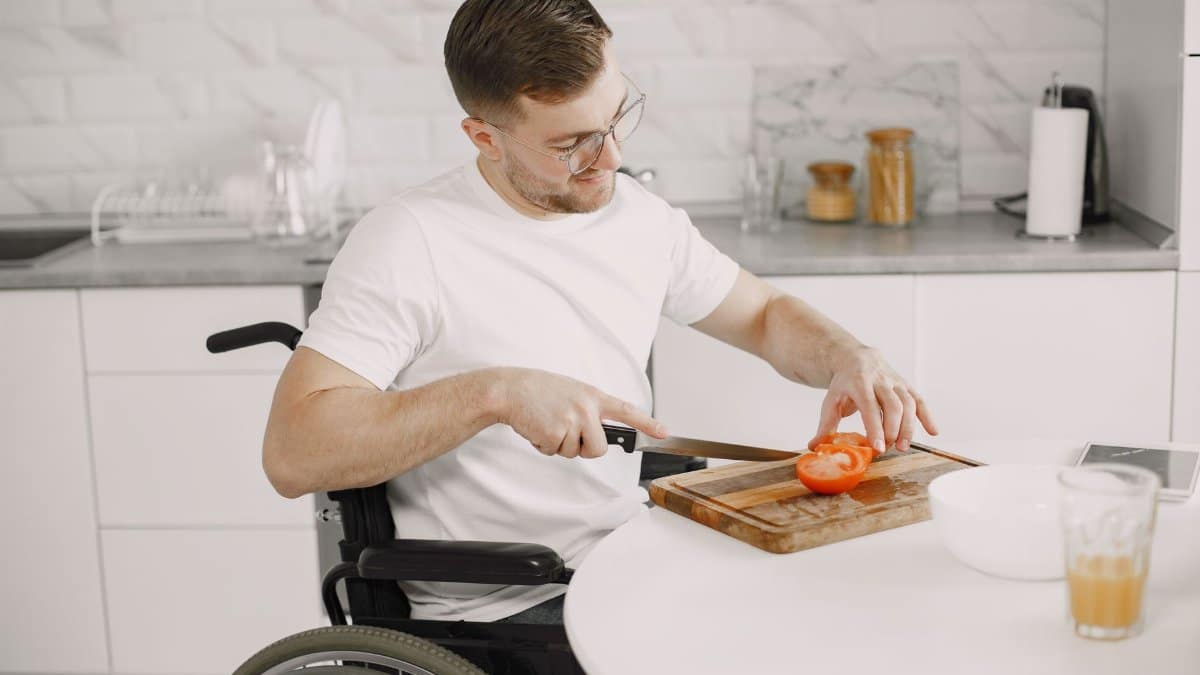 Man in a wheelchair slices a tomato on a wooden board in a modern kitchen setting.