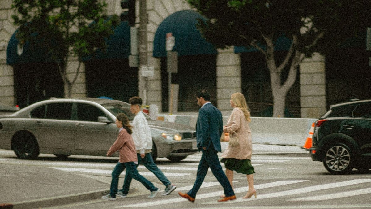 A family crosses a city street at a crosswalk surrounded by cars and buildings.