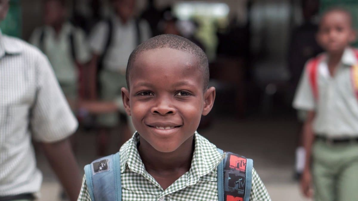 A cheerful schoolboy in a Haitian school uniform, carrying a backpack outdoors.