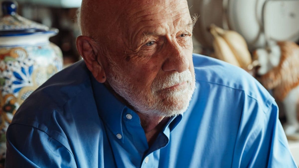 Elderly man with beard in a blue shirt, thoughtfully gazing indoors.
