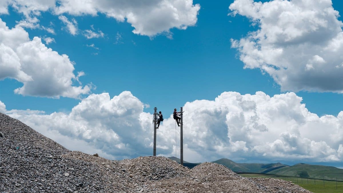 Two workers climb utility poles amid rocky terrain and vibrant clouds.