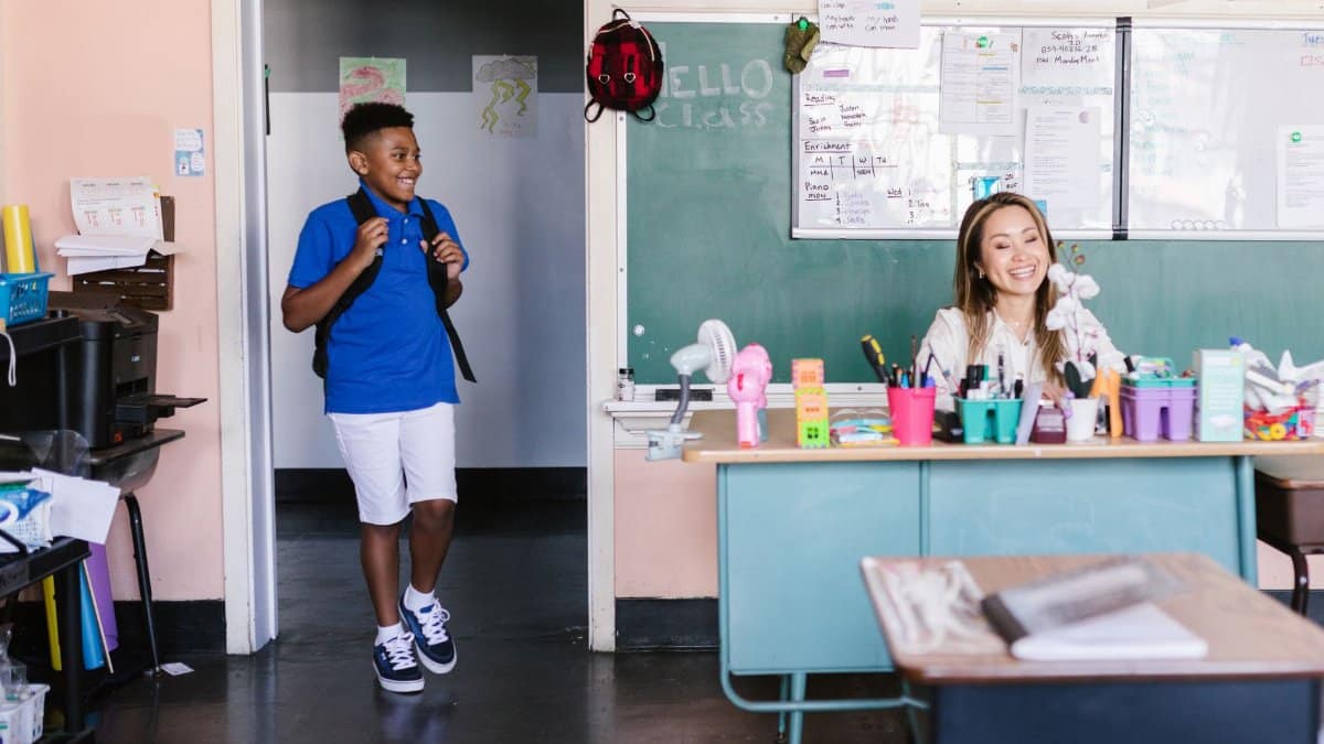 A cheerful student enters the classroom where a teacher is seated at a desk, ready for class.