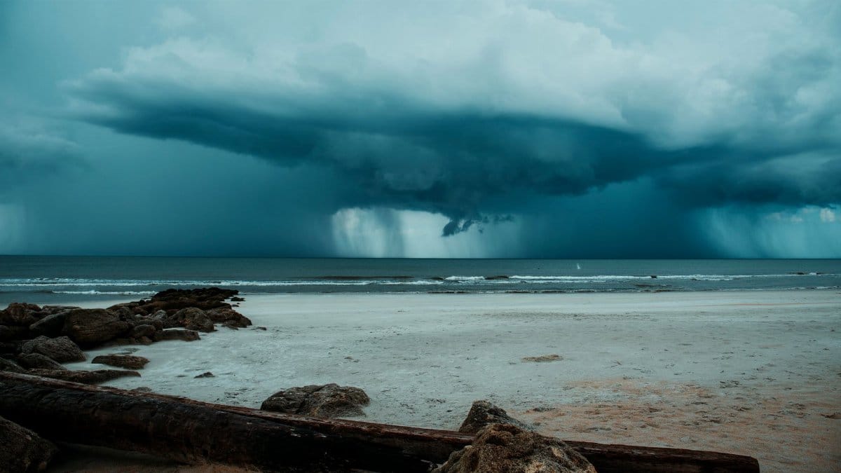Turbulent storm clouds loom over a quiet beach with driftwood and rocks in view.